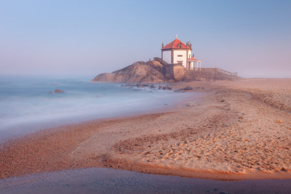 senhor da Pedra Chapel, a small white chapel perched on a rocky outcrop in Vila Nova de Gaia, Portugal. The chapel is surrounded by a sandy beach and the vast Atlantic Ocean. -